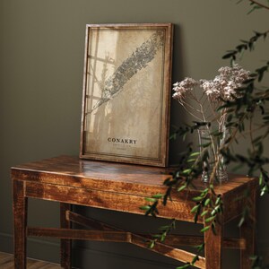 May include: A framed map print of Conakry, Guinea, with a vintage aesthetic. The map is in shades of brown and beige, set in a dark wooden frame. The print is displayed on a dark wooden table.