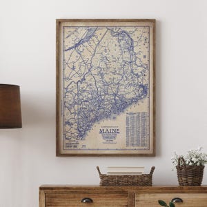 May include: A framed vintage map of Maine, featuring blue ink details on a beige background. The map is enclosed in a brown wooden frame and displays the state's roads, cities, and geographical features. The word "MAINE" is prominently displayed at the bottom.