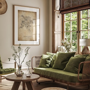 May include: A living room scene featuring a framed map with the text "YOUR CITY". A green velvet sofa with matching pillows sits near a wooden coffee table with vases of white flowers. A woven ottoman and a window with a bamboo shade are also visible.