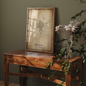 May include: A framed map of Monroe, Louisiana, in a vintage style. The map is printed on aged paper and set in a dark wood frame. The text "MONROE" is displayed at the bottom of the map. The framed map is displayed on a wooden table.