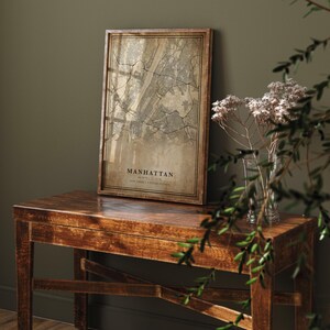 May include: A framed map of Manhattan, New York, in a vintage style. The map is in shades of brown and beige, with the city's streets and waterways clearly visible. The frame is made of dark wood, and the text "Manhattan" is printed at the bottom.