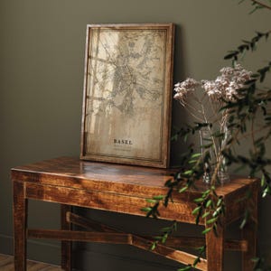 Peut inclure: Une carte encadrée de Bâle, Suisse, avec une esthétique vintage. La carte est dans des tons bruns et beiges, avec le nom de la ville et le pays imprimés en bas. Le cadre est en bois foncé. La carte est posée sur une table en bois.