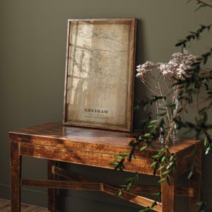 May include: A framed map print of Gresham, Oregon, in a vintage style. The map is in shades of brown and tan, with the city name and coordinates displayed. The frame is made of dark wood, and the print is displayed on a wooden table.