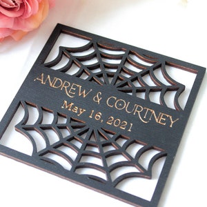 May include: A square black wooden plaque featuring a laser-cut spider web design. The center horizontal band displays the engraved text "ANDREW & COURTNEY" above the date "May 16, 2021" in a gold-toned font, with visible wood grain edges.