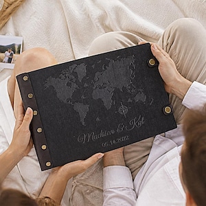 May include: A black leather-bound photo album with a world map design and the names "Madison & Kai" and the date "05.14.2022" embossed on the cover. The album is held by two people. The album has brass-colored corner protectors and binding posts.