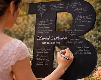 May include: A person signing a large, black wooden letter "B" guest book at a wedding. The letter is decorated with white calligraphy and the names "Daniel & Amber" and the date "X0-413-2023".
