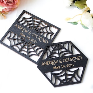 May include: Two black wooden coasters with laser-cut spider web patterns. One coaster is square and the other is hexagonal. Both feature the gold-colored text "ANDREW & COURTNEY" and "May 16, 2021" engraved across the center of the web design.