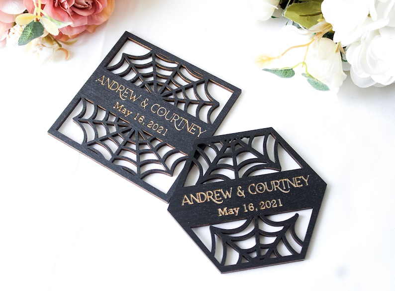 May include: Two black wooden coasters with laser-cut spider web patterns. One coaster is square and the other is hexagonal. Both feature the gold-colored text "ANDREW & COURTNEY" and "May 16, 2021" engraved across the center of the web design.