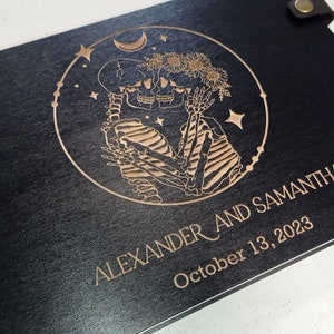 May include: A black wooden photo album with a brown leather spine and brass rivets. The front cover features a laser-engraved design of two skeletons holding hands, surrounded by stars and a crescent moon. The text "ALEXANDER AND SAMANTHA" and "October 13, 2023" is also engraved on the cover.