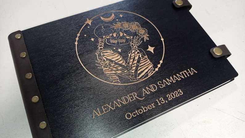 May include: A black wooden photo album with a brown leather spine and brass rivets. The front cover features a laser-engraved design of two skeletons holding hands, surrounded by stars and a crescent moon. The text "ALEXANDER AND SAMANTHA" and "October 13, 2023" is also engraved on the cover.