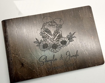 May include: A dark brown wooden guest book with a laser engraved design of two skeletons kissing surrounded by flowers. The text "Shayla & Jondi" is engraved below the skeletons.