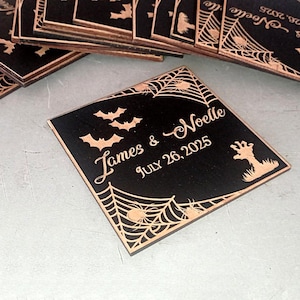May include: A stack of square black coasters featuring laser-engraved copper-toned details including spider webs, bats, a zombie hand, and the text James & Noelle JULY 26, 2025. The design is consistent across all pieces in the collection.