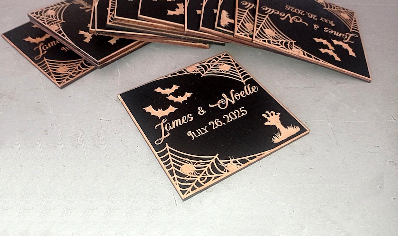 May include: A stack of square black coasters featuring laser-engraved copper-toned details including spider webs, bats, a zombie hand, and the text James & Noelle JULY 26, 2025. The design is consistent across all pieces in the collection.