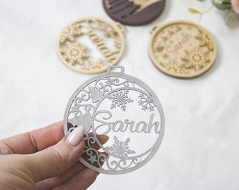 May include: A silver-colored, laser-cut Christmas ornament with the name "Sarah" and snowflake details. Other wooden ornaments with names and winter designs are in the background. The ornament is held by a person.