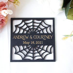 May include: A square black wooden plaque featuring a laser-cut spider web design. The center displays the gold-colored text ANDREW & COURTNEY and the date May 16, 2021. The plaque is surrounded by pink and white artificial flowers.