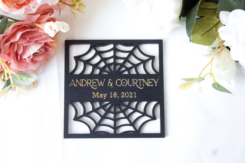 May include: A square black wooden plaque featuring a laser-cut spider web design. The center displays the gold-colored text ANDREW & COURTNEY and the date May 16, 2021. The plaque is surrounded by pink and white artificial flowers.