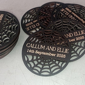 May include: A stack and cluster of circular black coasters featuring a laser-cut spider web pattern. Each coaster is engraved with the text CALLUM AND ELLIE and the date 14th September 2025 in a light tan color across a central horizontal bar.