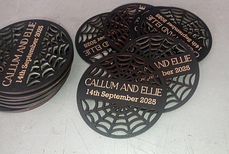 May include: A stack and cluster of circular black coasters featuring a laser-cut spider web pattern. Each coaster is engraved with the text CALLUM AND ELLIE and the date 14th September 2025 in a light tan color across a central horizontal bar.