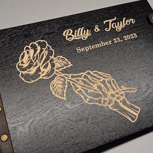 May include: A black wooden guest book with a brown leather spine and brass rivets. The cover features an engraved design of a rose held by a skeletal hand. The text "Billy & Taylor" and "September 23, 2023" is also engraved on the cover.