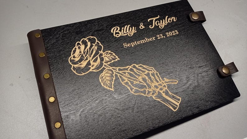 May include: A black wooden guest book with a brown leather spine and brass rivets. The cover features an engraved design of a rose held by a skeletal hand. The text "Billy & Taylor" and "September 23, 2023" is also engraved on the cover.