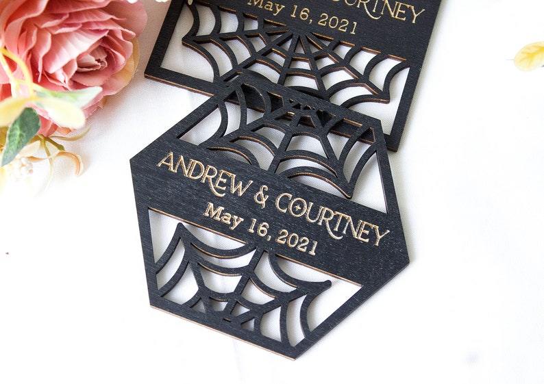 May include: Two black laser-cut wooden ornaments featuring a spider web design. Each piece is engraved with the text ANDREW & COURTNEY and the date May 16, 2021 in gold lettering. The ornaments have a geometric shape with intricate cutouts.