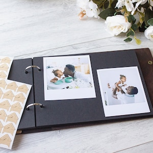 May include: A black photo album with a brown leather cover and metal rings. The album is open to reveal two photos of a father and daughter. The photos are printed on white paper and have a white border. The album also has a set of brown corner stickers.