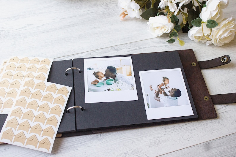May include: A black photo album with a brown leather cover and metal rings. The album is open to reveal two photos of a father and daughter. The photos are printed on white paper and have a white border. The album also has a set of brown corner stickers.