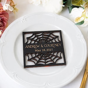 May include: A square, dark-stained wooden coaster featuring a laser-cut spider web pattern. The center displays the gold-colored text "ANDREW & COURTNEY" above the date "May 16, 2021" in a clean, sans-serif font, centered on a white plate.