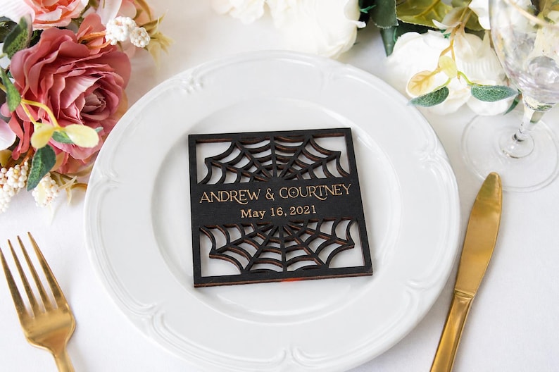 May include: A square, dark-stained wooden coaster featuring a laser-cut spider web pattern. The center displays the gold-colored text "ANDREW & COURTNEY" above the date "May 16, 2021" in a clean, sans-serif font, centered on a white plate.