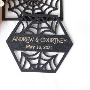 May include: A black hexagonal wooden coaster featuring a laser-cut spider web pattern. The center displays the gold-colored text ANDREW & COURTNEY and the date May 16, 2021. The design includes intricate web cutouts above and below the text.