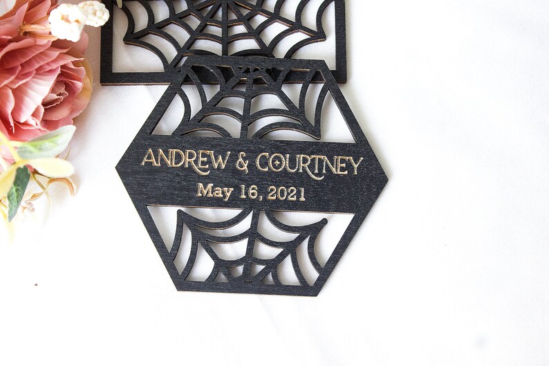 May include: A black hexagonal wooden coaster featuring a laser-cut spider web pattern. The center displays the gold-colored text ANDREW & COURTNEY and the date May 16, 2021. The design includes intricate web cutouts above and below the text.