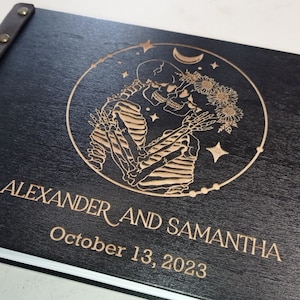 May include: Black wooden photo album with a leather strap and brass accents. The cover features an engraved design of two skeletons embracing, surrounded by stars and a crescent moon. The text "ALEXANDER AND SAMANTHA October 13, 2023" is engraved below the design.