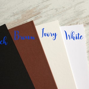 May include: Four samples of cardstock in different colors: black, brown, ivory, and white. The colors are labeled in blue cursive writing.