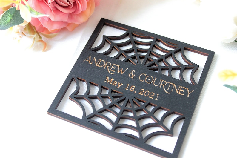 May include: A square black wooden plaque featuring a laser-cut spider web design. The center horizontal band displays the engraved text "ANDREW & COURTNEY" above the date "May 16, 2021" in a gold-toned font, with visible wood grain edges.