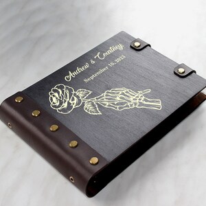 May include: A dark brown leather-bound photo album with gold accents. The cover features a gold-colored rose and a skeleton hand holding the stem. The album is personalized with the text "Andrew & Courtney" and "September 10, 2023".