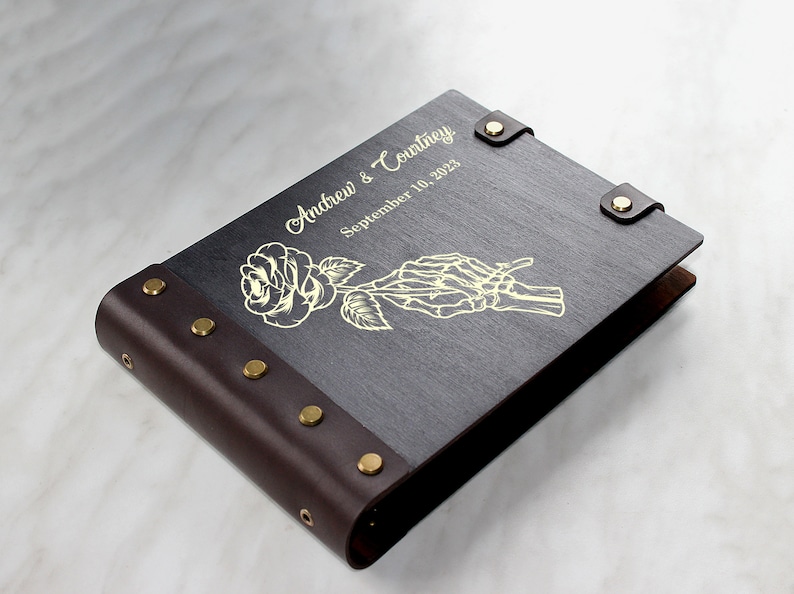 May include: A dark brown leather-bound photo album with gold accents. The cover features a gold-colored rose and a skeleton hand holding the stem. The album is personalized with the text "Andrew & Courtney" and "September 10, 2023".