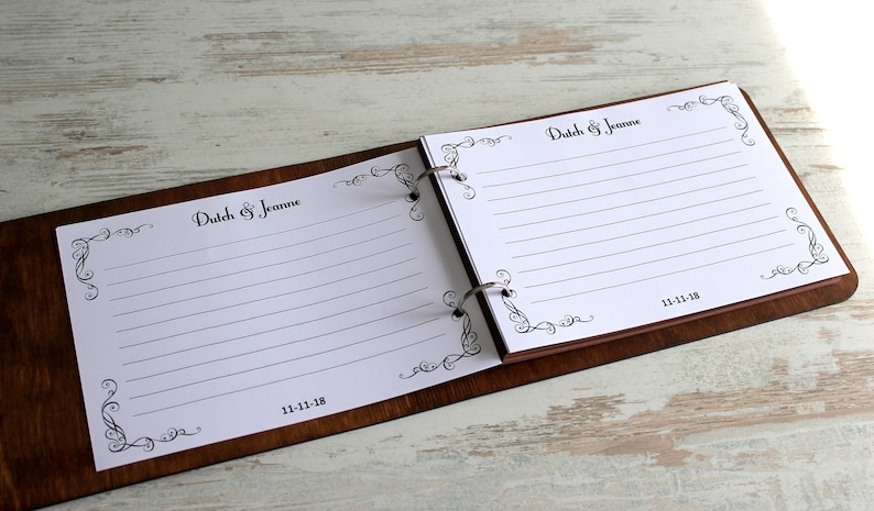 May include: A brown wooden recipe book with a white paper insert. The book is open to show the lined pages. The book has a decorative border with the text "Dutch & Jeanne" and the date "11-11-18".