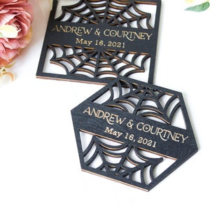 May include: Two black wooden coasters with laser-cut spider web patterns. Each coaster features the engraved gold text "ANDREW & COURTNEY" and "May 16, 2021". One coaster is square-shaped, and the other is hexagonal, both displaying intricate web designs.