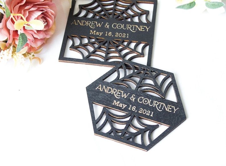 May include: Two black wooden coasters with laser-cut spider web patterns. Each coaster features the engraved gold text "ANDREW & COURTNEY" and "May 16, 2021". One coaster is square-shaped, and the other is hexagonal, both displaying intricate web designs.