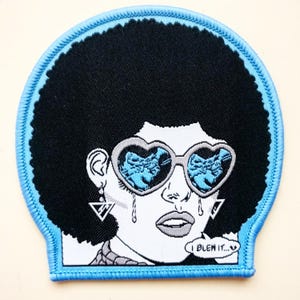 May include: A black and white embroidered patch of a woman with an afro wearing heart-shaped sunglasses and crying. The text "I BLEW IT..." is in a speech bubble. The patch has a light blue border.