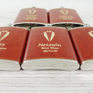May include: Six brown leather-covered hip flasks with silver trim. Each flask has a gold engraved tuxedo with a bow tie and personalised text. The text includes names, titles, and dates.