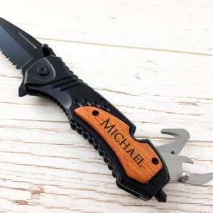 Op de afbeelding: Een zwart vouwmes met een houten handvat gegraveerd met de naam "MICHAEL". Het mes heeft een gekarteld blad en een flesopener.