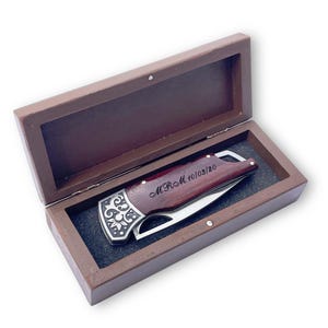 May include: A brown wooden box with a hinged lid, housing a folding knife. The knife features a dark wood handle with the engraved text "MRM 10/03/20" and a decorative silver-coloured metal bolster. The box is brown.