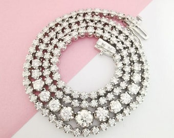 May include: A silver diamond tennis necklace with a row of large round diamonds in the center and smaller diamonds on either side.