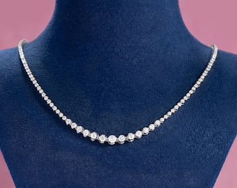 May include: A delicate silver necklace with numerous round, sparkling gemstones. The necklace is displayed on a blue velvet surface, with a pink background. The gemstones increase in size towards the center of the necklace.