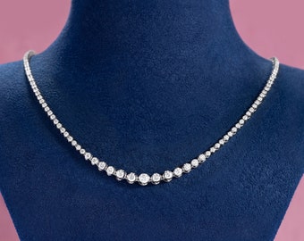 May include: A delicate diamond necklace with a single row of round diamonds set in a silver or white gold setting. The necklace is displayed on a blue velvet background.