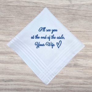 Pode incluir: Lenço branco com texto bordado em azul: "I'll see you at the end of the aisle, Your Wife" e um símbolo de coração. O lenço tem formato quadrado com um design de borda listrada.