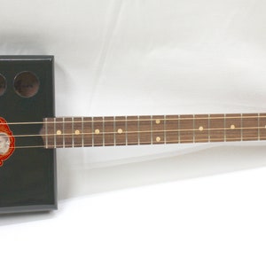 May include: A black cigar box guitar with a gold and red label that says "Calabash". The guitar has a wooden neck with a brown fretboard and gold frets.