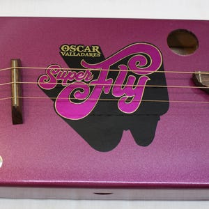 Puede incluir: Una guitarra hecha a mano con una caja de cigarros con un acabado púrpura metálico. La guitarra presenta las palabras "Super Fly" en un diseño rosa y negro, con el nombre "Oscar Valladares" arriba. Tiene cuerdas y un diapasón de madera.