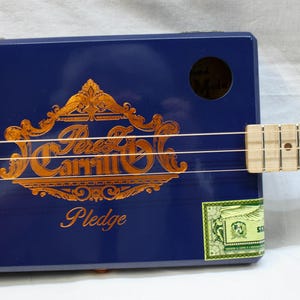 Könnte beinhalten: Eine handgefertigte Zigarrenkistengitarre mit einem marineblauen Korpus und einem hellen Holzgriffbrett. Die Gitarre trägt den Text "Perez Carrillo Pledge" in goldfarbenen Buchstaben. Auf der Seite befindet sich ein kleines grün-weißes Etikett.